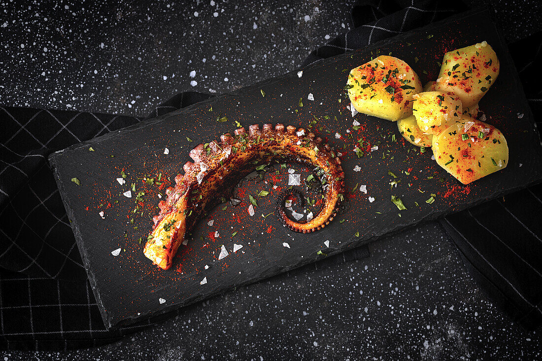 From above of fried octopus tentacle and pieces of potato served with spices on black board on table