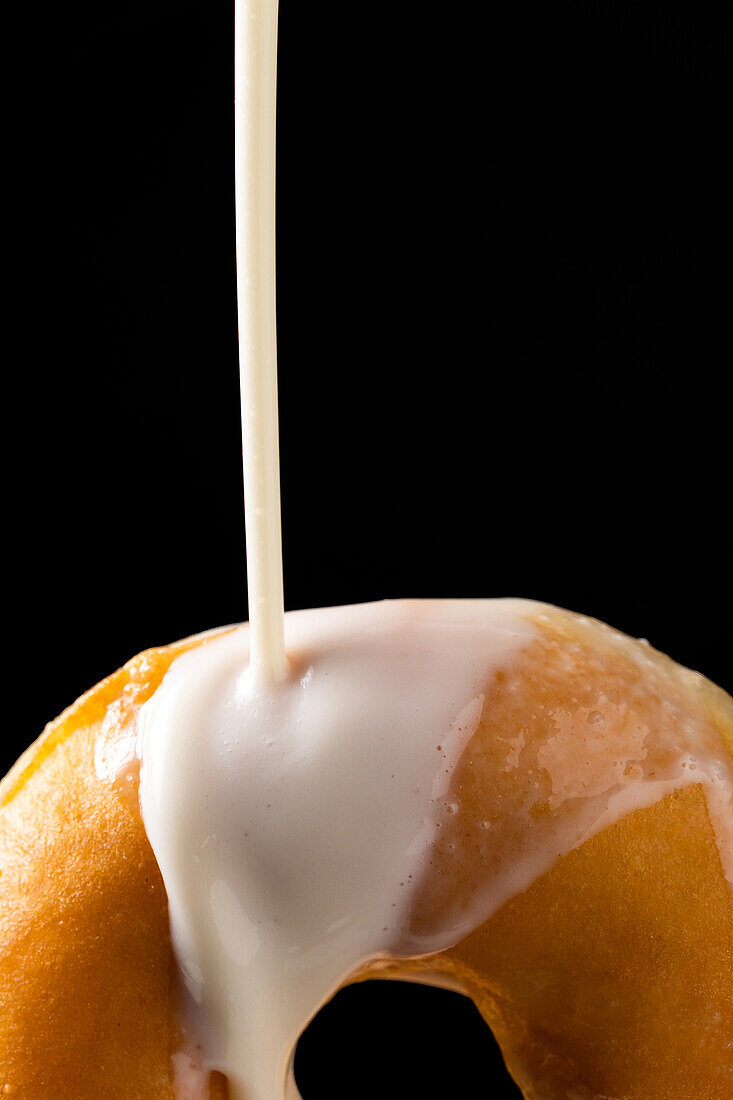 Anonyme Person gießt weißen Zuckerguss Milch auf leckeren süßen Donut vor schwarzem Hintergrund