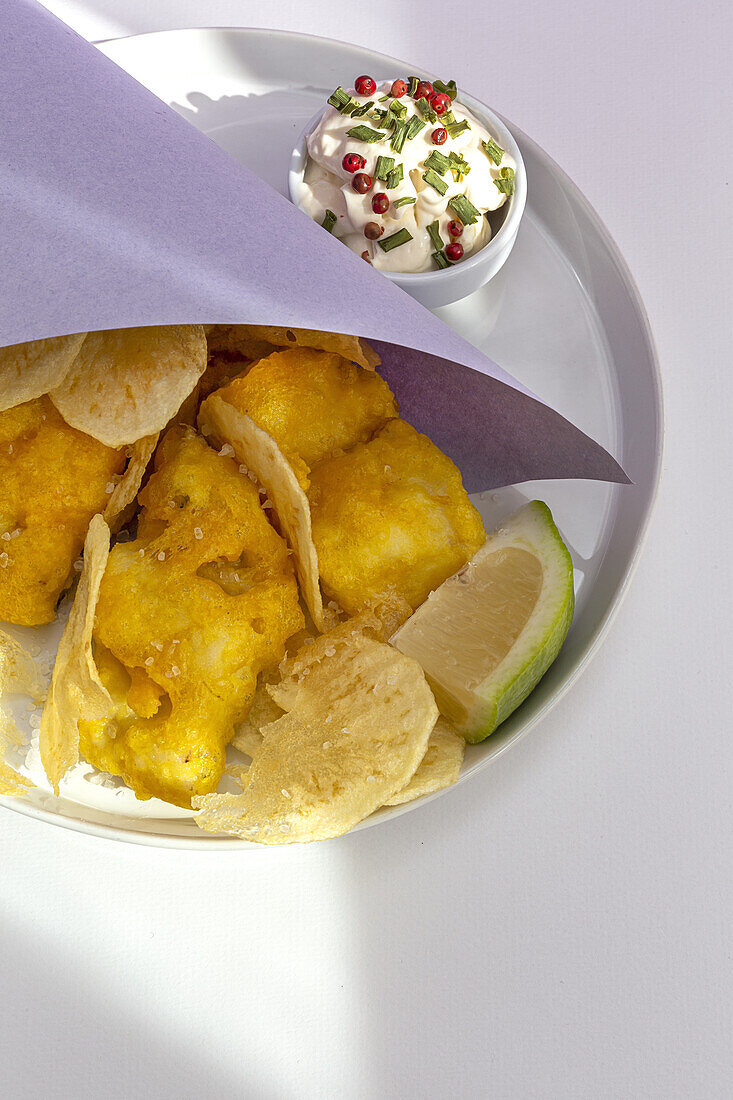 Appetizing fried chips and fish in paper wrapping served on plate with sauce on table with bright sunlight in cafe