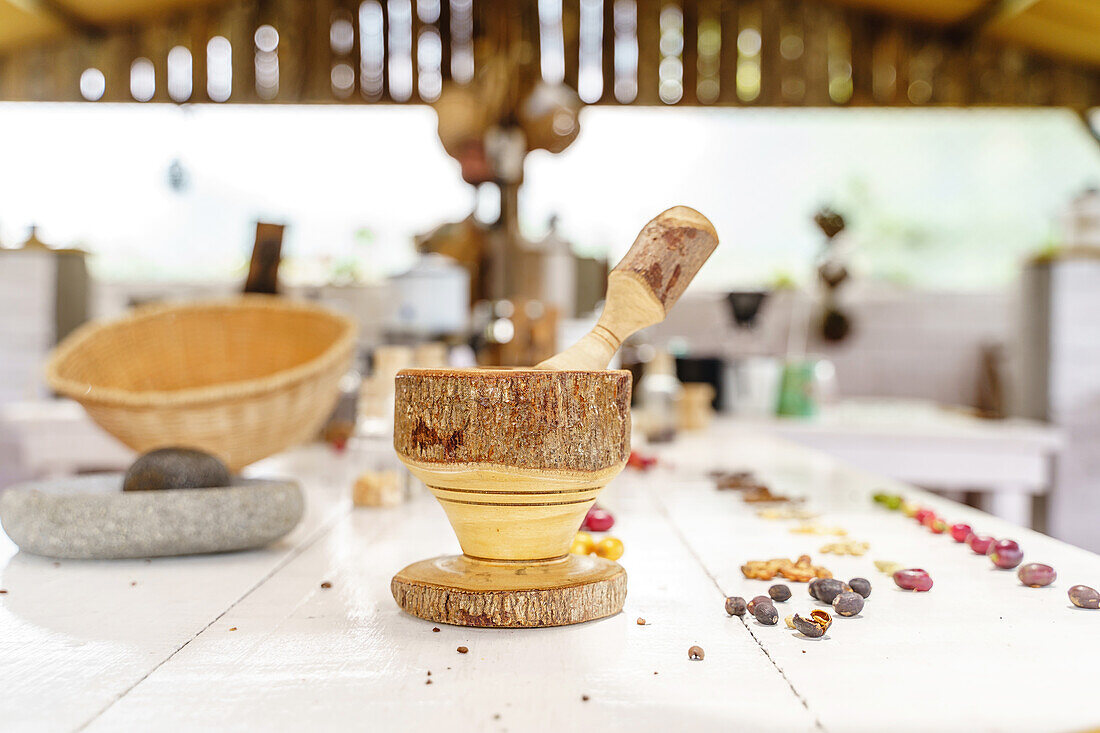 Wooden mortar and pestle placed against coffee berries and beans on table on blurred background of stone plate and basket