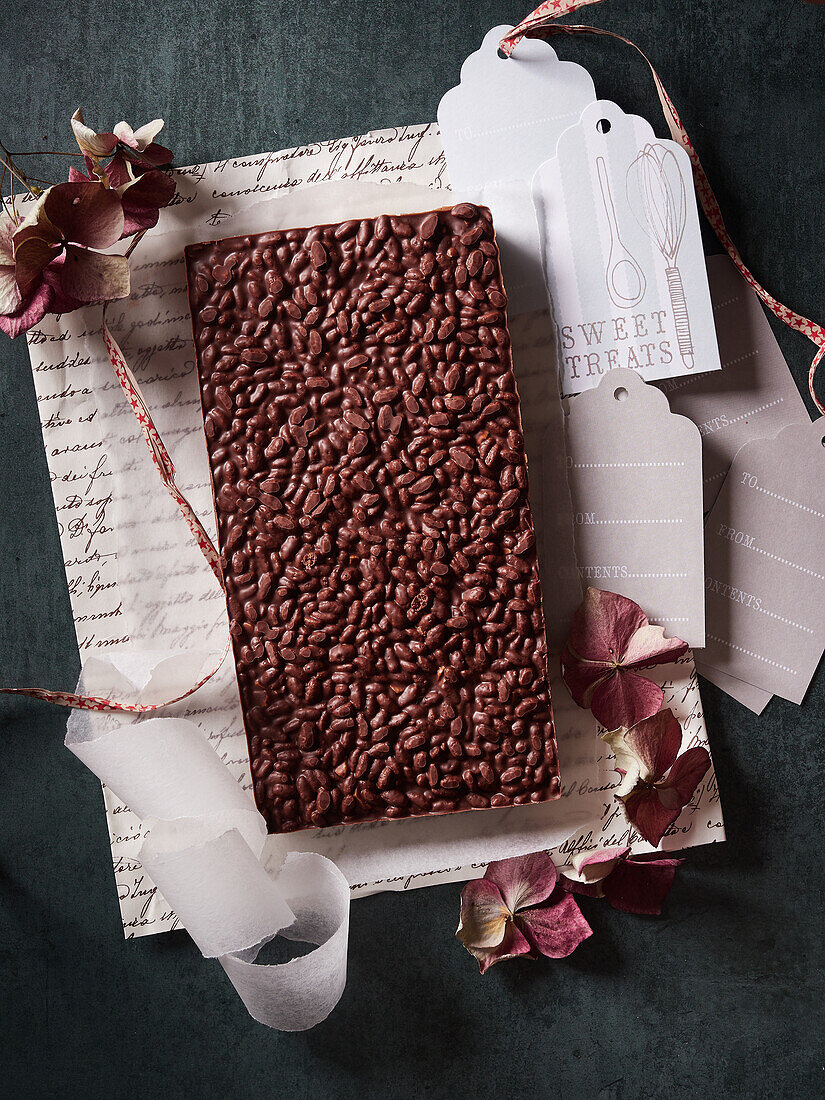 Draufsicht auf süße leckere Reis-Crispy-Schokoladen-Crunch-Riegel auf Geschenkpapier mit getrockneten Blumen und Tags auf dunklem Hintergrund