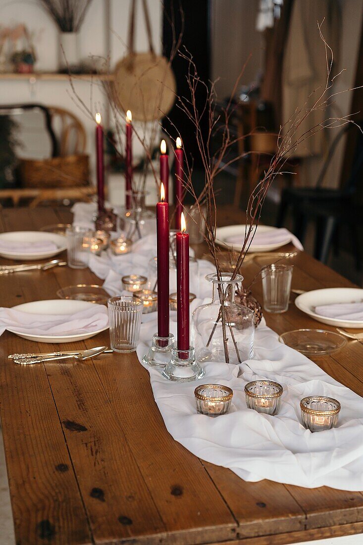 Weißes Tischtuch und Teller auf einem festlich gedeckten Tisch mit brennenden Kerzen und trockenen Baumzweigen