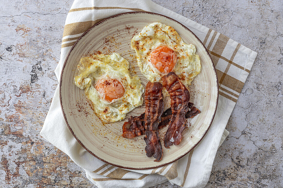 Overhead view of tasty sunny side up eggs with fried bacon strips on plate above towel