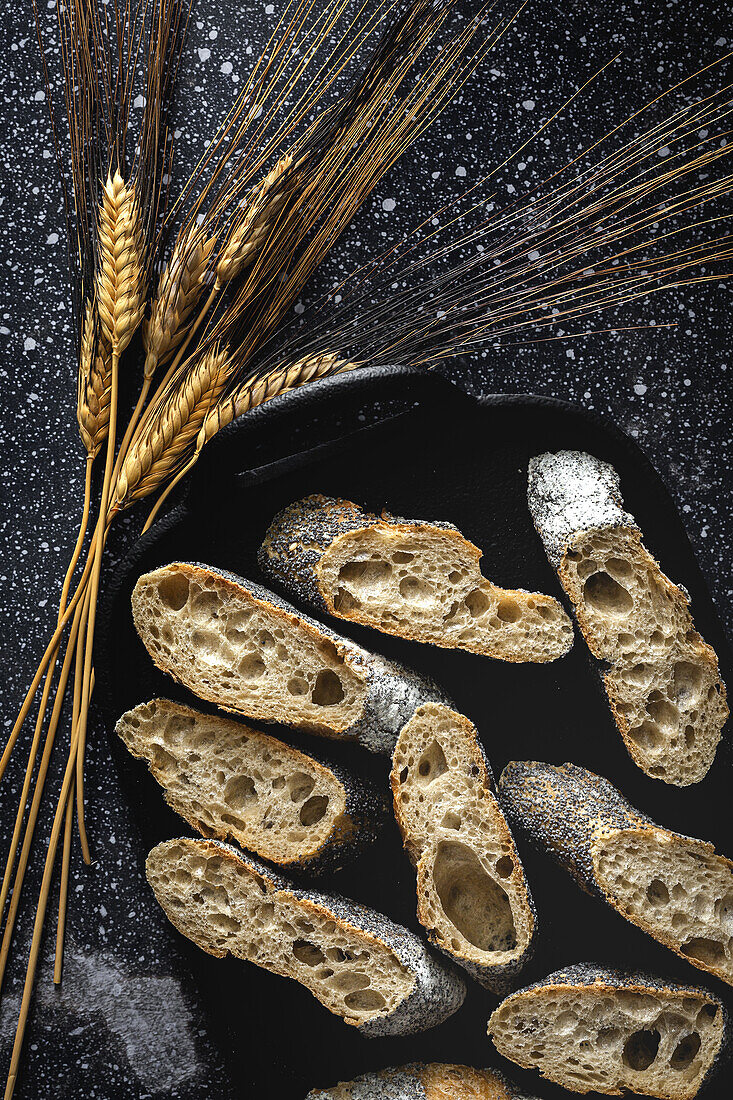 From above of appetizing crusty bread near wheat spikes and dark fabric on table