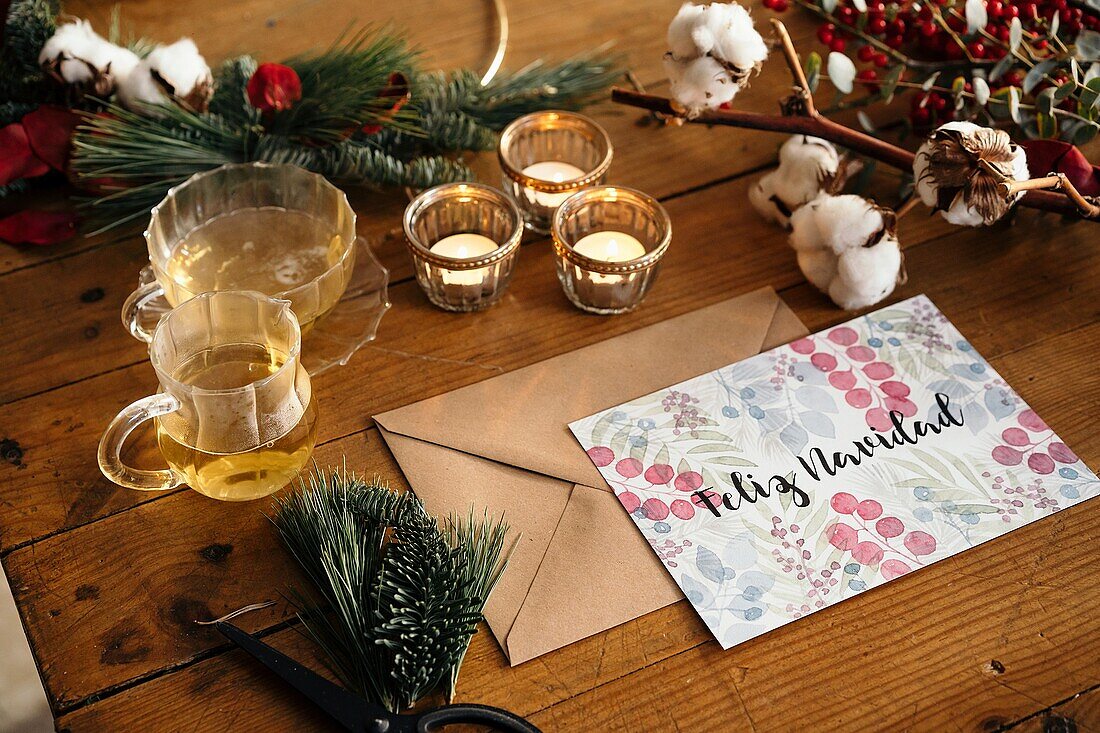 Draufsicht auf eine weihnachtliche Komposition mit einer bunten Postkarte mit der Aufschrift Feliz Navidad in der Nähe von brennenden Kerzen und Teetassen auf einem Holztisch, der mit bunten Pflanzenzweigen dekoriert ist
