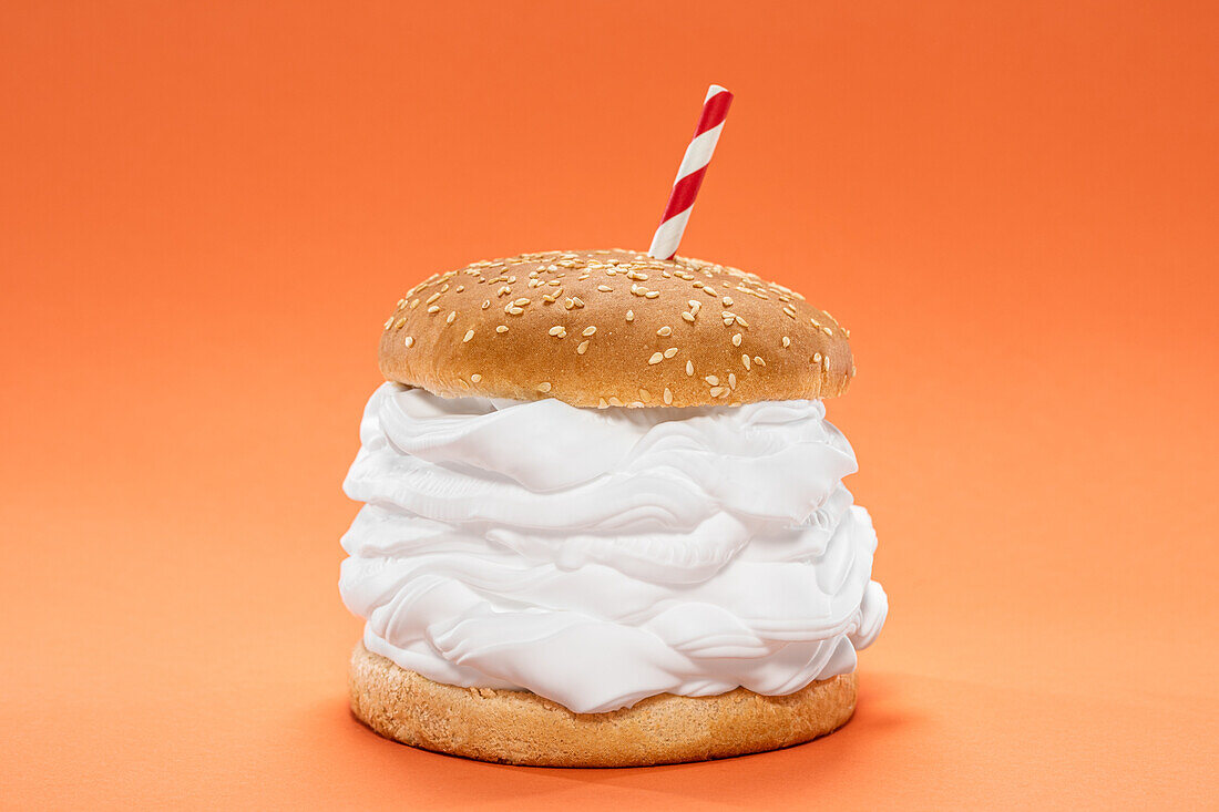 Halves of burger bun with sesame and sweet whipped cream and straw placed against orange background
