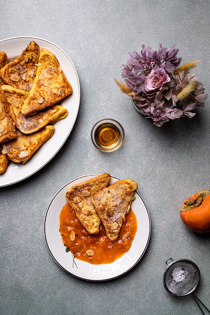 Top view of white plate with delicious French toasts with jam placed near jar with honey and fresh persimmons on gray table