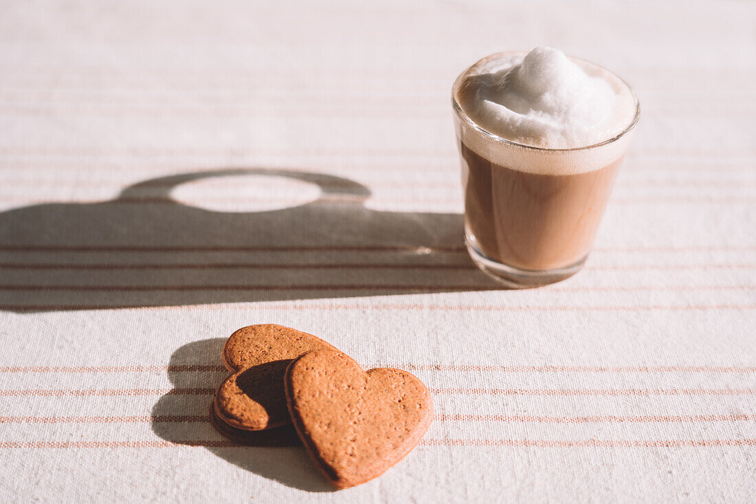 From above glass of foamy latte macchiato served on table with heart shaped cookies on sunny day in kitchen