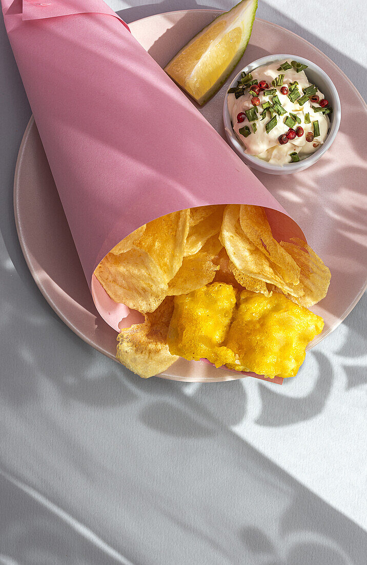 Appetitlich gebratene Pommes frites und Fisch in Papierverpackung auf einem Teller mit Soße auf einem Tisch mit hellem Sonnenlicht in einem Café serviert