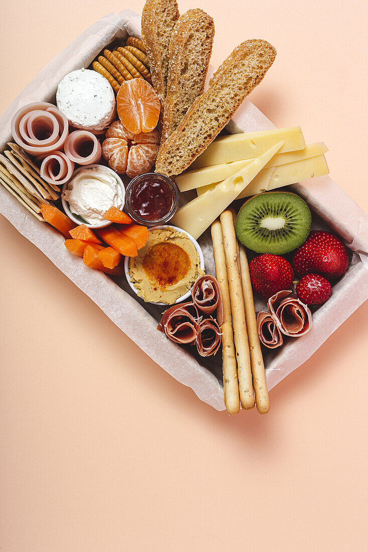 From above brunch box with assorted sliced meats various types of cheese and crispbreads arranged near ripe cup kiwi sweet strawberries and peeled mandarin near jam in glass jar on colorful pastel background
