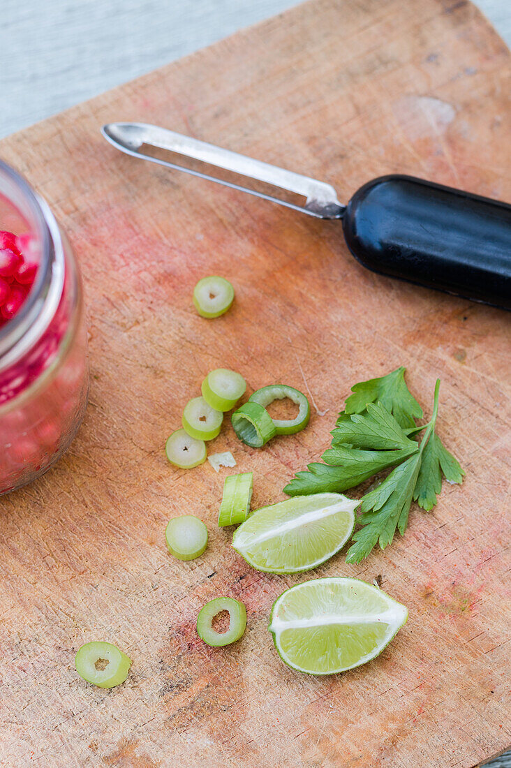 From above of green herbs and lime pieces placed near vegetable peeler and glass jar with pomegranate seeds on wooden chopping board