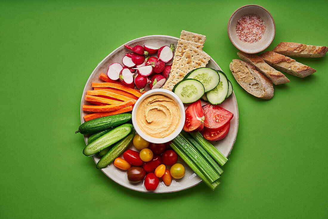 From above of appetizing fresh raw cut cucumbers with carrots and radish served on white table with assorted tomato and hummus and placed on green table with pieces of bread