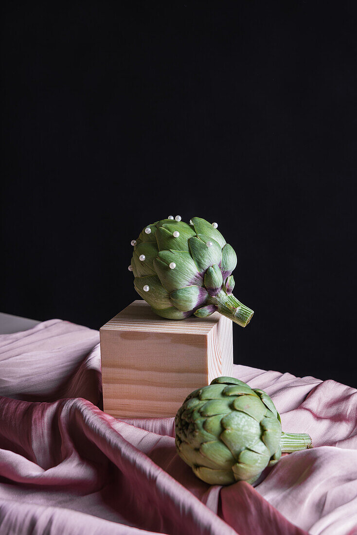 Ripe artichokes placed on wooden cube on table with silk fabric against black background
