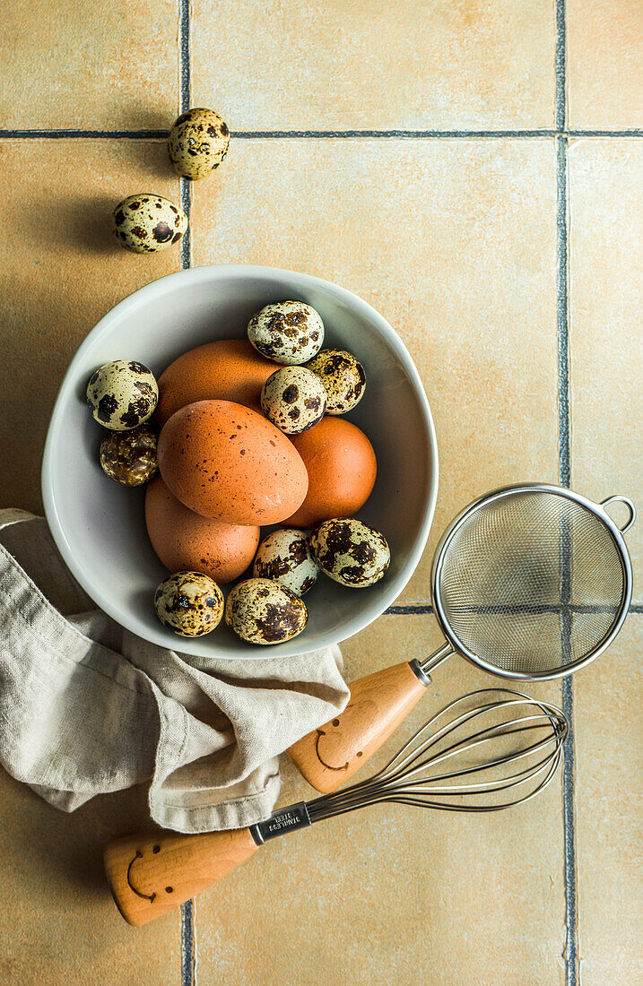 From above ceramic bowl with raw quail and chicken eggs near kitchen utensils on tiled table