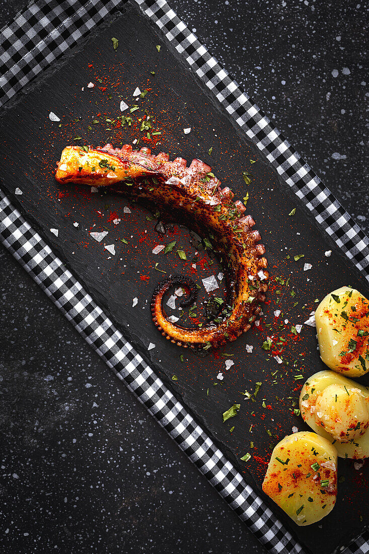 Top view of fried octopus tentacle and pieces of potato served with spices on black board on table