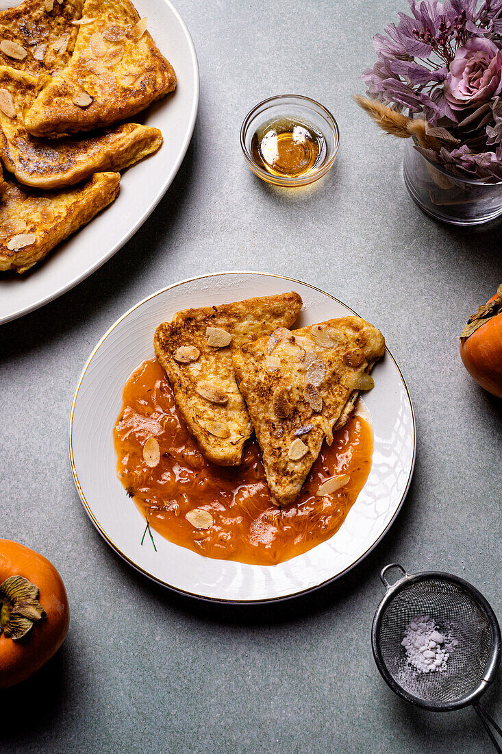 Top view of white plate with delicious French toasts with jam placed near jar with honey and fresh persimmons on gray table