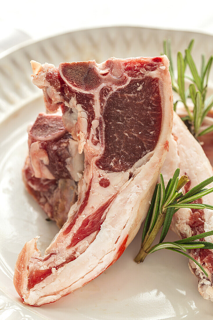 From above of uncooked lamb chops served on white ceramic plate with fresh green rosemary on table in light kitchen