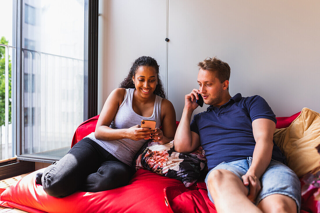 Cheerful young diverse couple in casual clothes resting on red couch using smartphone