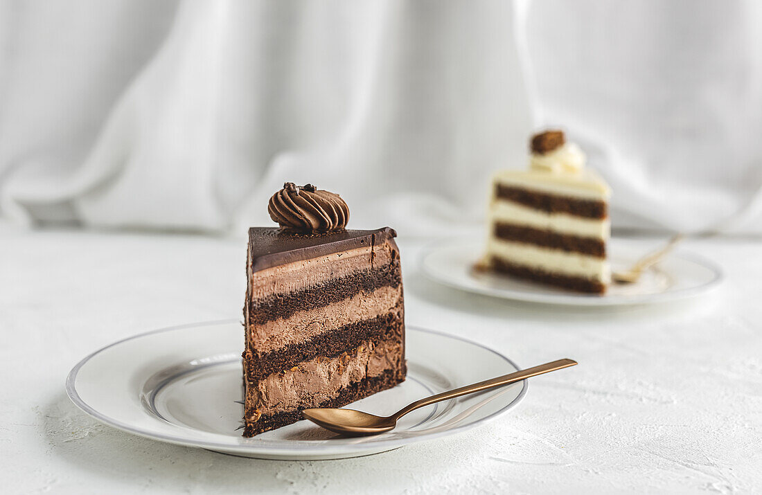 Piece of yummy sweet chocolate cake and white cake with glaze and whipped cream served on ceramic plate with golden spoon on white tablecloth