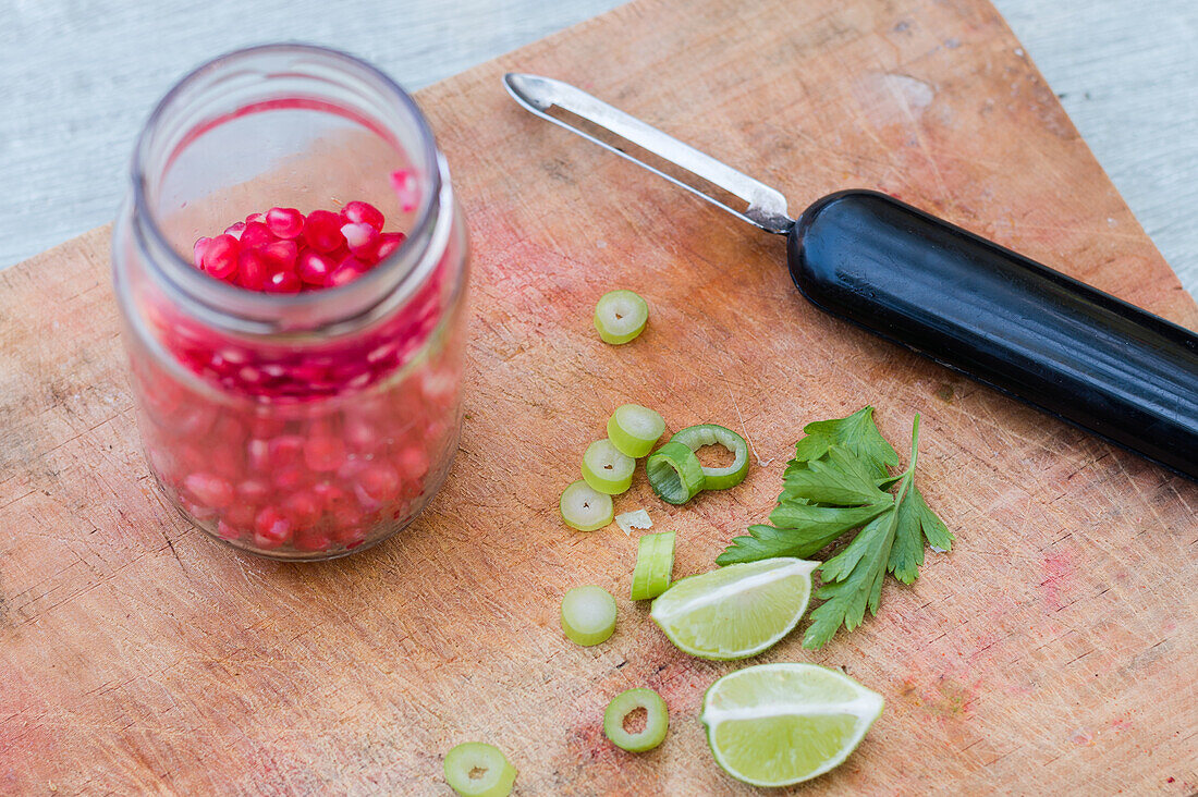 From above of green herbs and lime pieces placed near vegetable peeler and glass jar with pomegranate seeds on wooden chopping board