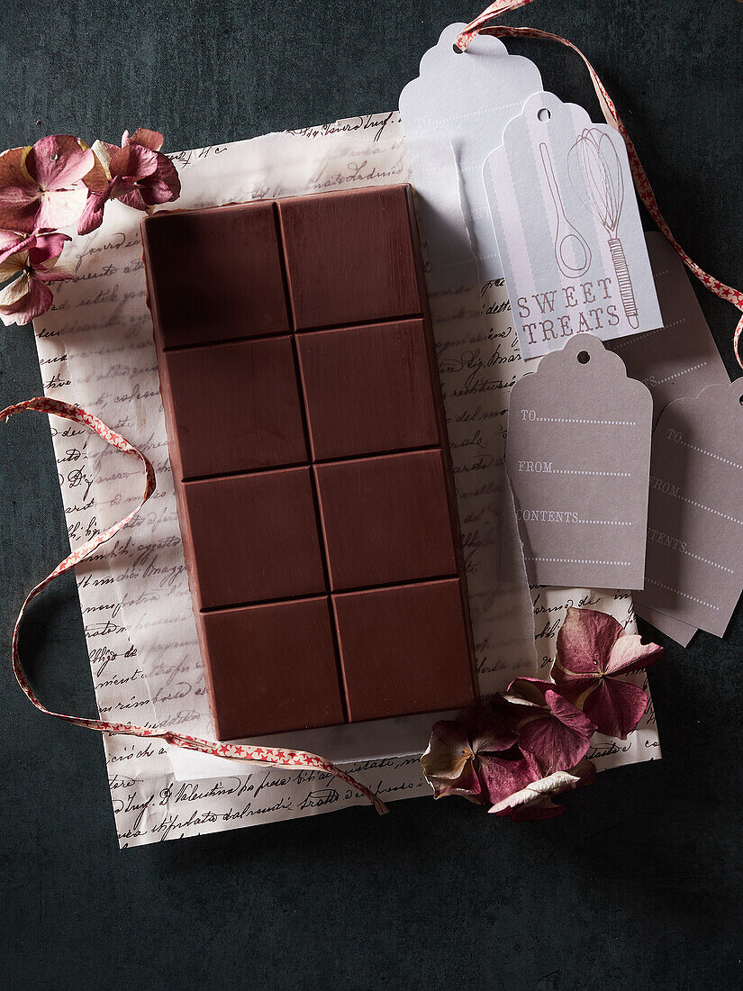 Draufsicht auf eine leckere süße Schokoladentafel auf Geschenkpapier mit getrockneten Blumen und Anhängern auf dunklem Hintergrund im Zimmer