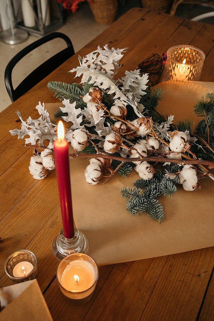 Festlicher Weihnachtsstrauß mit Baumwoll- und Tannenzweigen auf einem Holztisch mit Kerzen im Zimmer