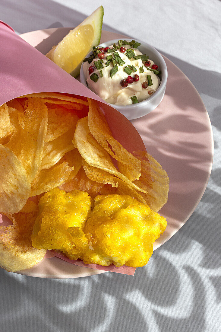 Appetitlich gebratene Pommes frites und Fisch in Papierverpackung auf einem Teller mit Soße auf einem Tisch mit hellem Sonnenlicht in einem Café serviert