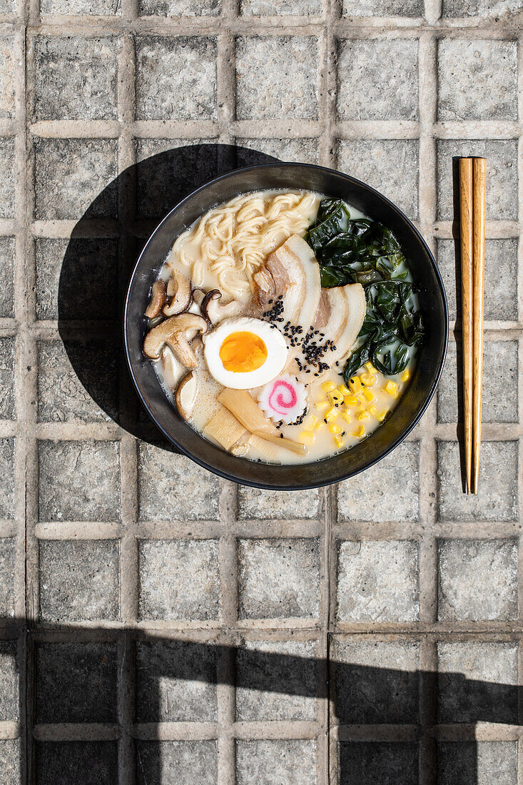 Top view of appetizing Japanese ramen with mushrooms and egg served on paved sidewalk with chopsticks on sunny street in city