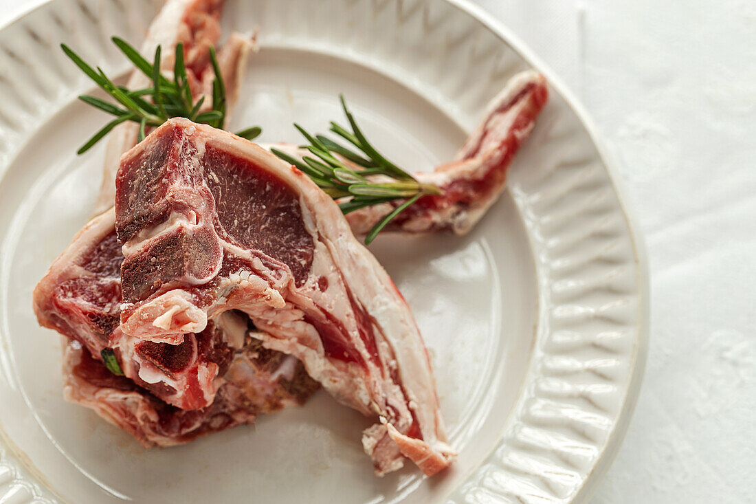 From above of uncooked lamb chops served on white ceramic plate with fresh green rosemary on table in light kitchen