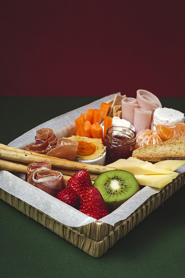Brunchbox mit verschiedenen Wurst- und Käsesorten und Knäckebrot, arrangiert neben reifen Kiwi, süßen Erdbeeren und geschälten Mandarinen, neben Marmelade in einem Glas auf buntem Hintergrund