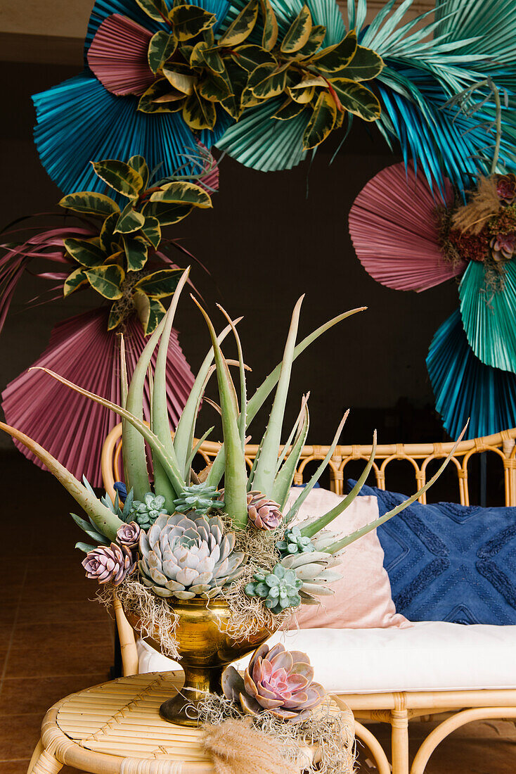 Elegante Vase mit einem Arrangement aus verschiedenen Sukkulenten auf einem kleinen Bambustisch in der Nähe eines bequemen Sofas auf der Terrasse, dekoriert mit kreativen, bunten, trockenen Palmenblättern