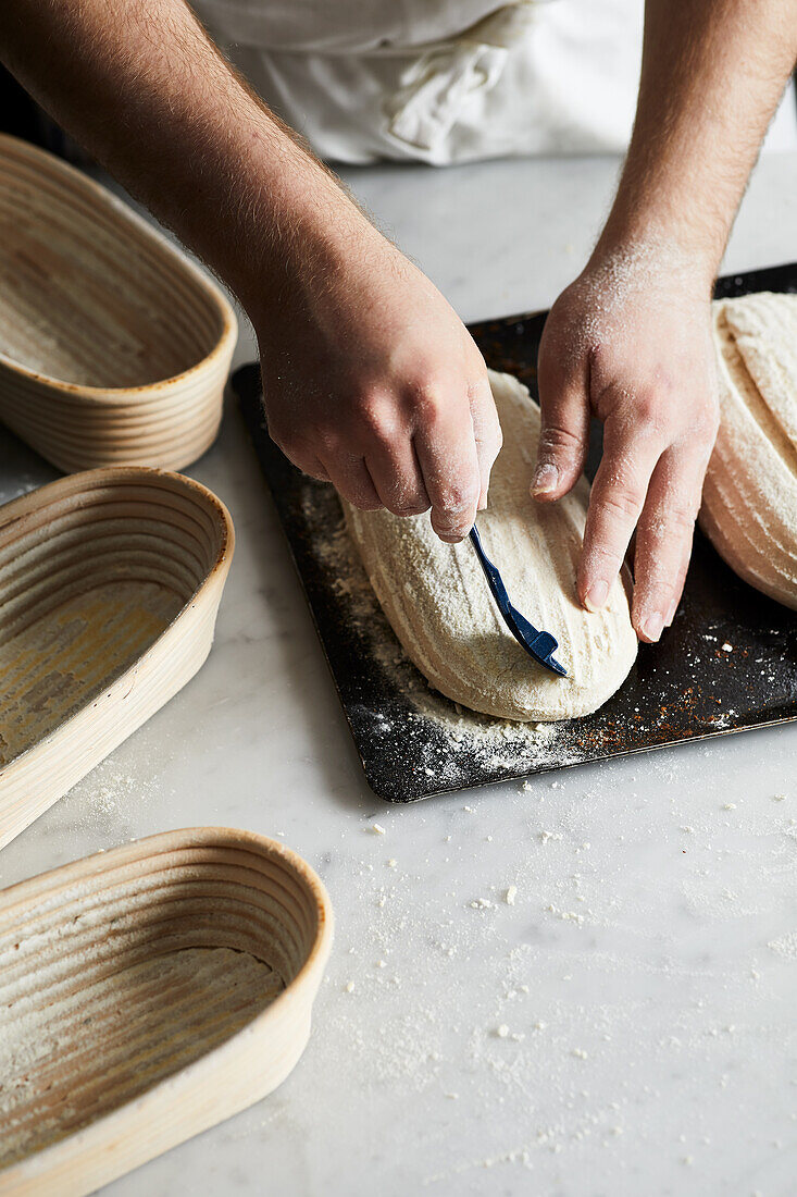 Anonymer Bäcker in Schürze mit Klinge ritzt rohen Sauerteig Laib auf Tisch während der Vorbereitung Brot in der Küche