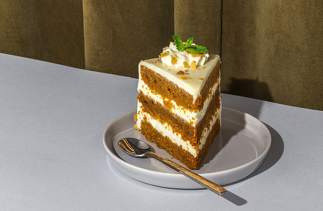Slice of tasty sweet carrot sponge cake with cream decorated with mint leaf served on plate with spoon on table in light room with curtain on the background