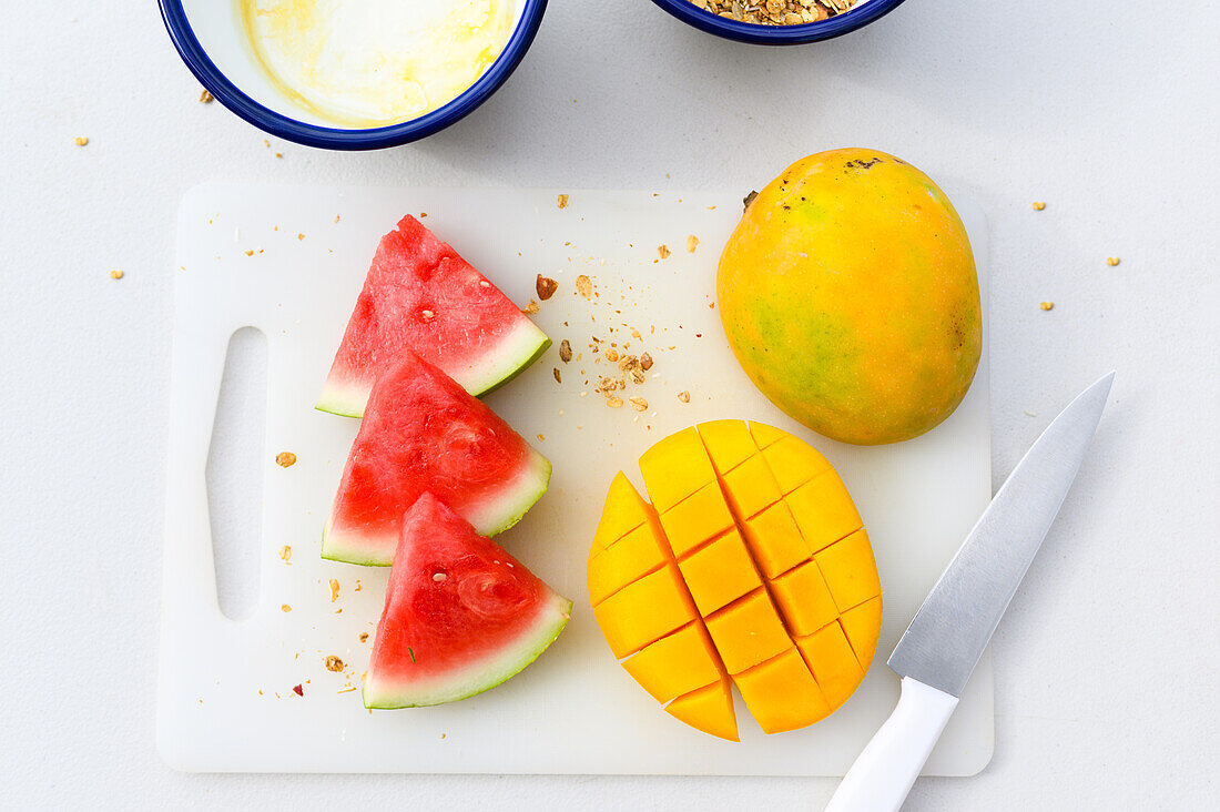 Draufsicht auf frische, in Scheiben geschnittene Wassermelone und orangefarbene, saftige, halbierte Mango, die mit einem Messer auf ein weißes Schneidebrett gelegt wird