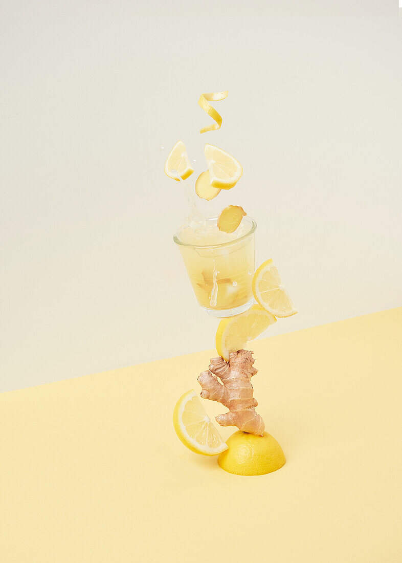 Slices of sour lemon falling into glass with ginger root and refreshing drink against two color background in light studio