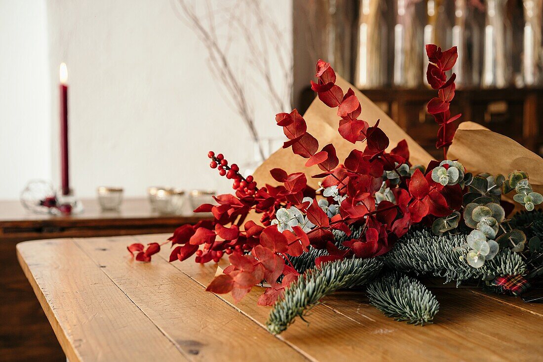 Festlicher, stilvoller, dekorativer Weihnachtsstrauß mit Eukalyptuszweigen und leuchtend roten Zweigen mit Beeren auf einem Holztisch im Zimmer
