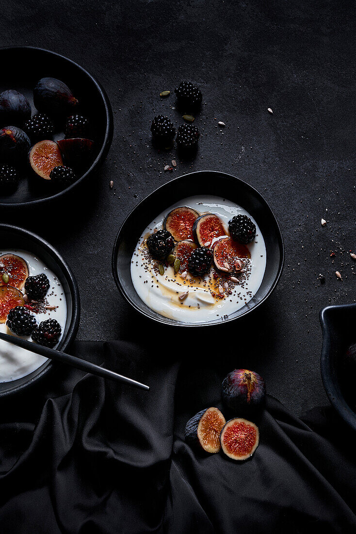 Appetitanregendes gesundes Frühstück bestehend aus Joghurt, frischen Feigen und Brombeeren, bestreut mit Samen und Nüssen auf einem schwarzen Tisch von oben