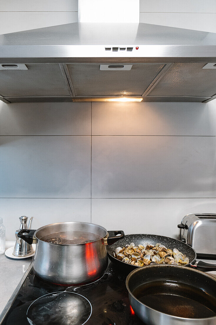 Clams frying in pan on electric cooker with pot and frying pan in light kitchen with extract hood at home