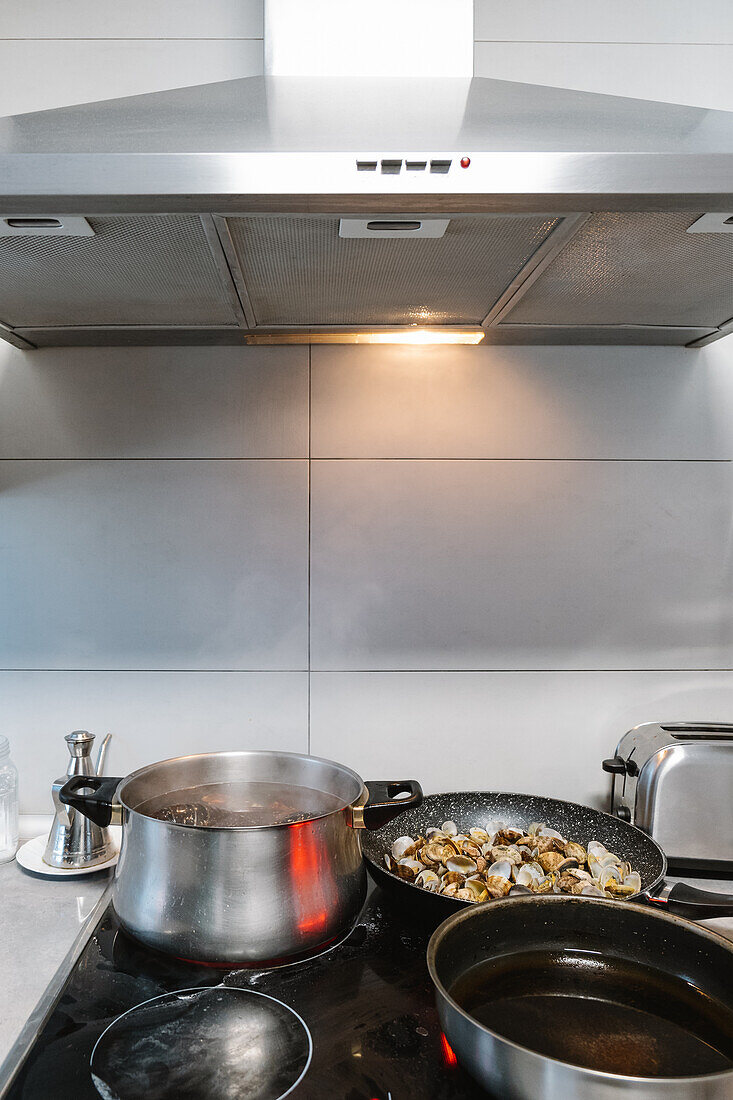 Muscheln braten in Pfanne auf Elektroherd mit Topf und Pfanne in heller Küche mit Abzugshaube zu Hause