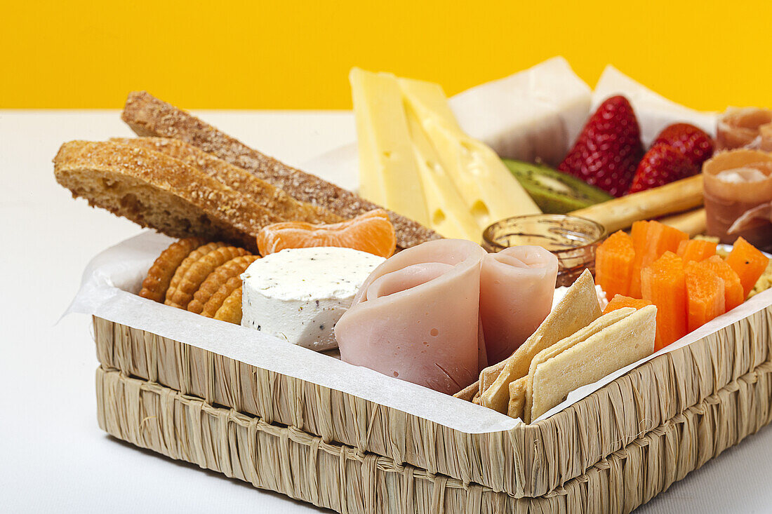 Brunchbox mit verschiedenen Wurst- und Käsesorten und Knäckebrot, arrangiert neben reifen Kiwi, süßen Erdbeeren und geschälten Mandarinen, neben Marmelade in einem Glas auf buntem Hintergrund