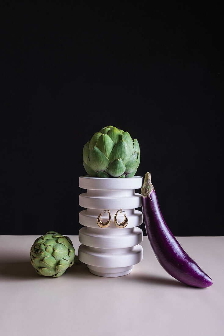 Ripe green artichokes and eggplant with precious accessories hanging on stand placed on table against black background