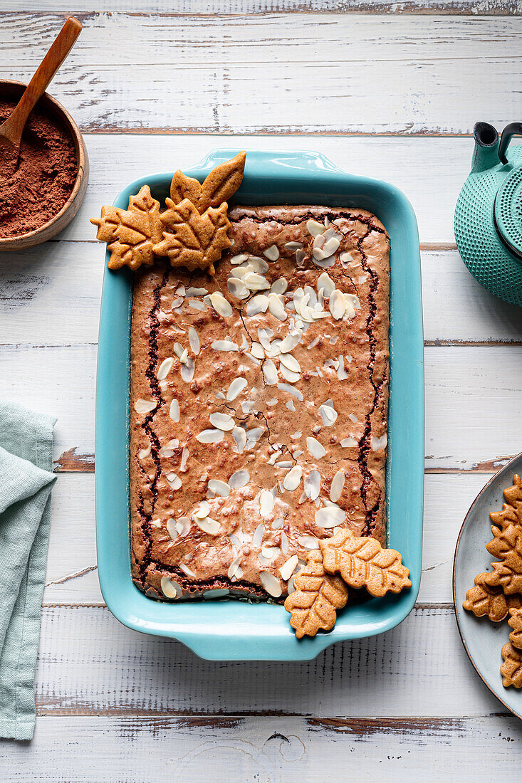 Draufsicht auf einen leckeren gebackenen Brownie mit Mandelblättchen in einem Backblech mit Keksen auf einem Holztisch mit Kakaopulver