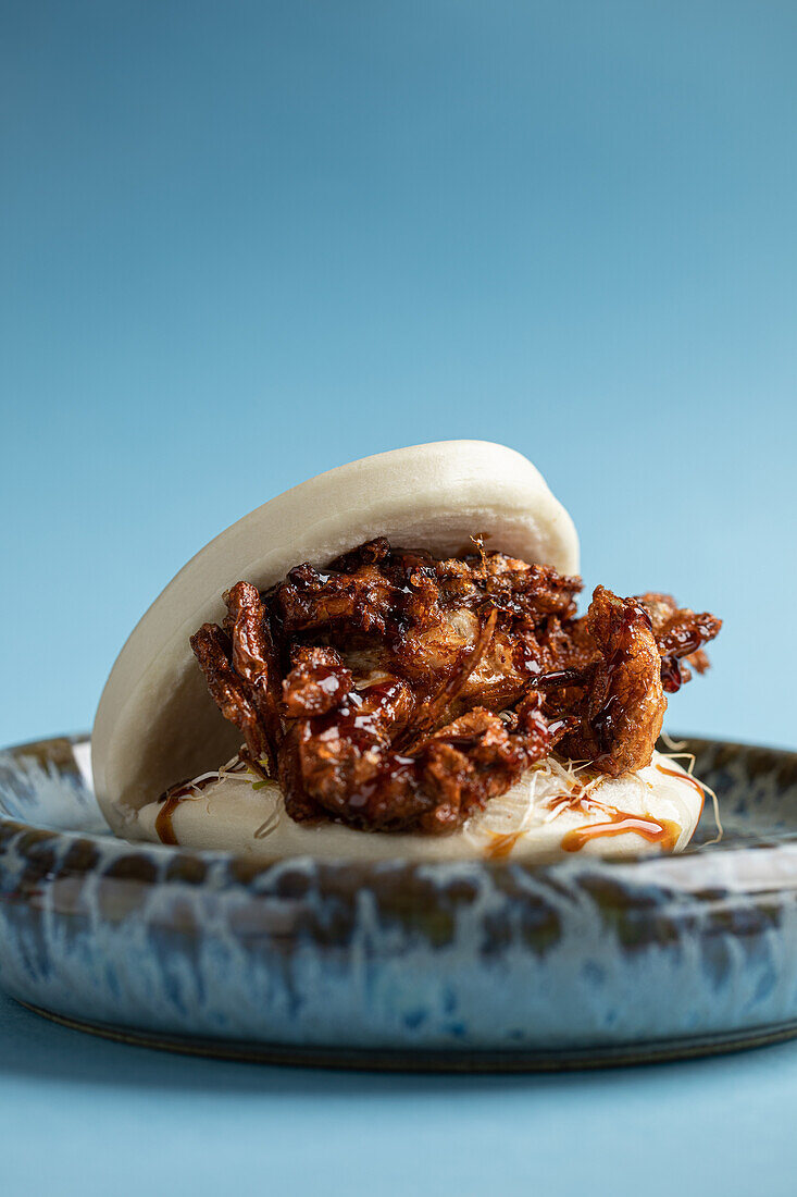 Delicious Japanese pan bao bun with fried soft shell crab served on ceramic plate against blue background in light studio