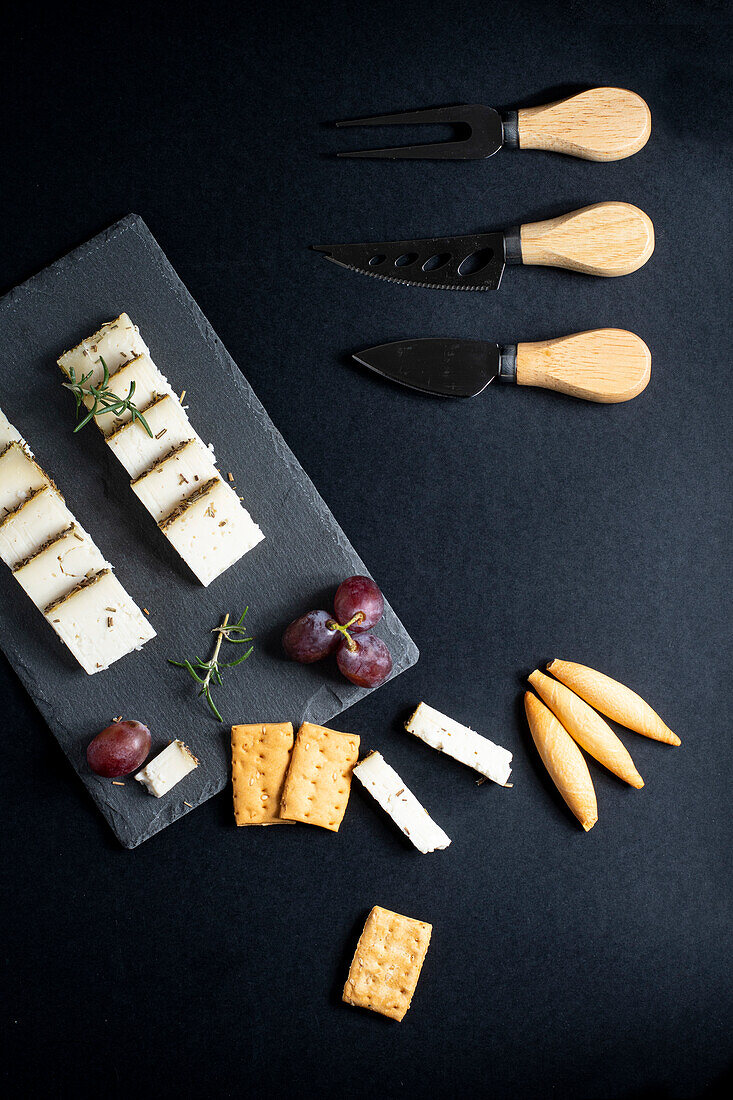 Top view of assorted delicious cheese arranged on black board with ripe grapes and crackers placed near knifes on dark surface