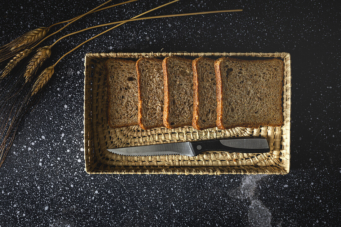 Top view of fresh homemade rye bread near knife in wicker basket and wheat spikes on table