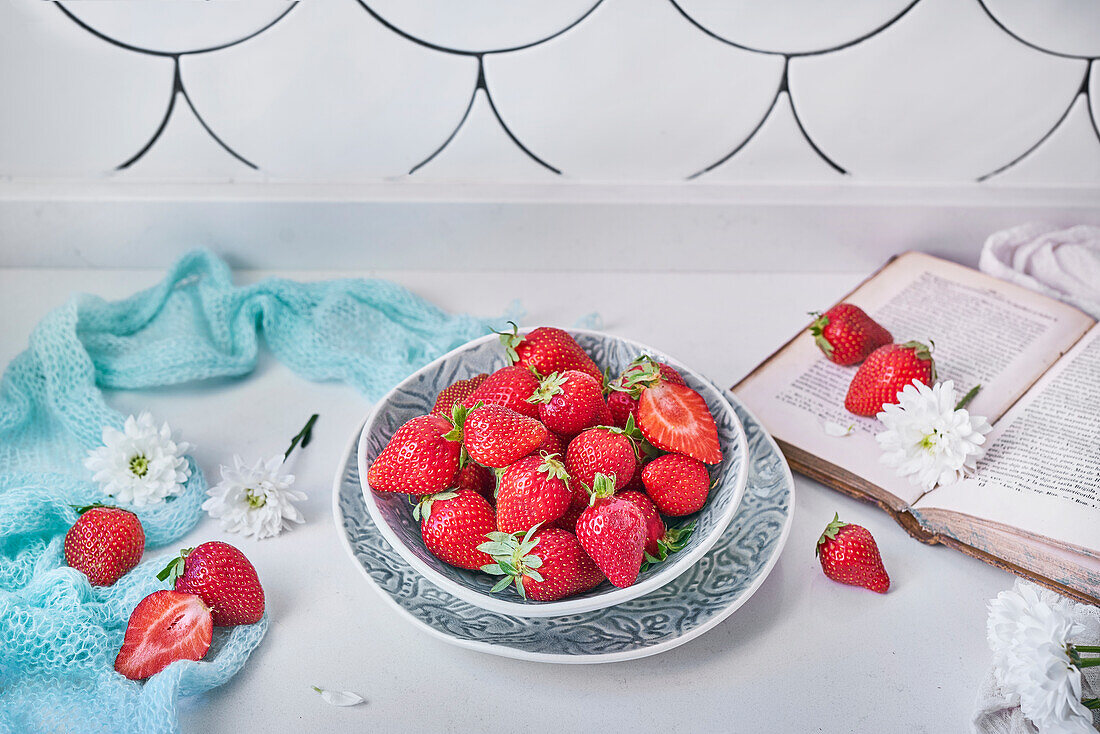 From above bowl of ripe strawberries placed on counter near fresh white flowers and opened book in daytime at home