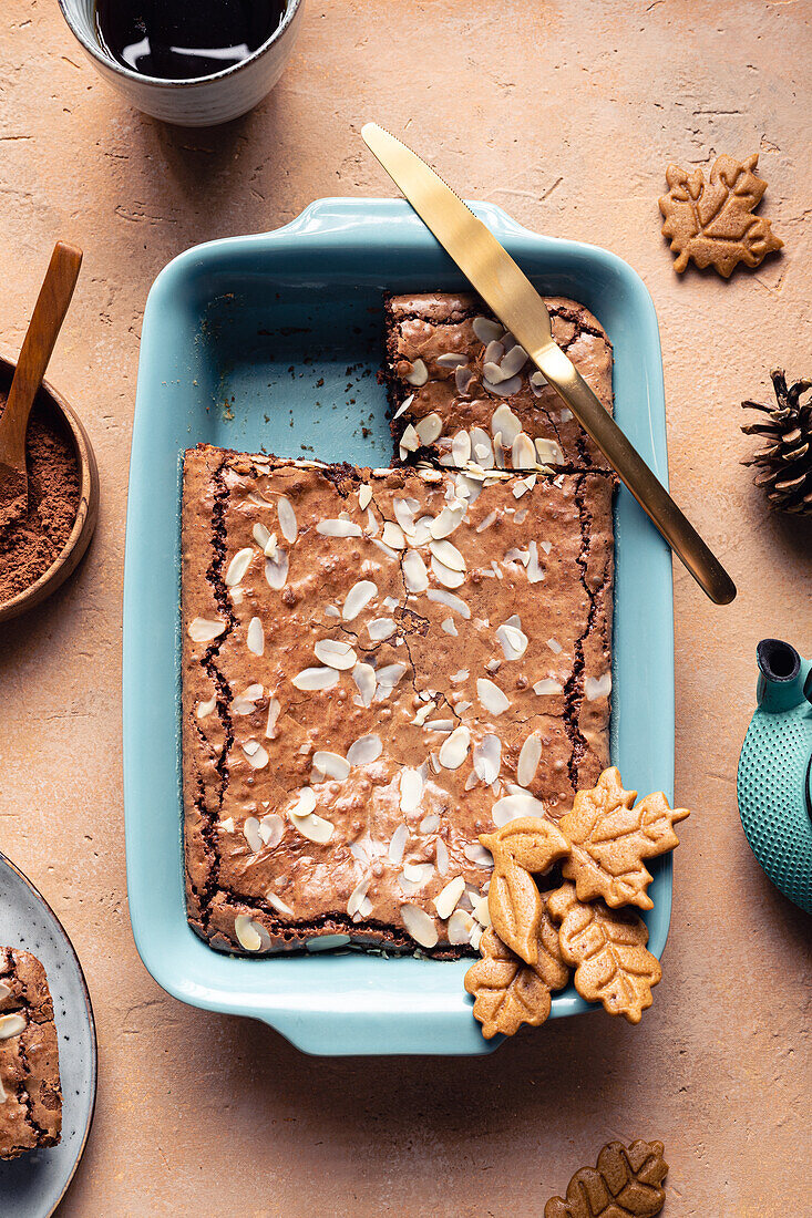 Top view of tasty baked brownie with almond flakes in baking tray with cookies placed on table with cocoa powder