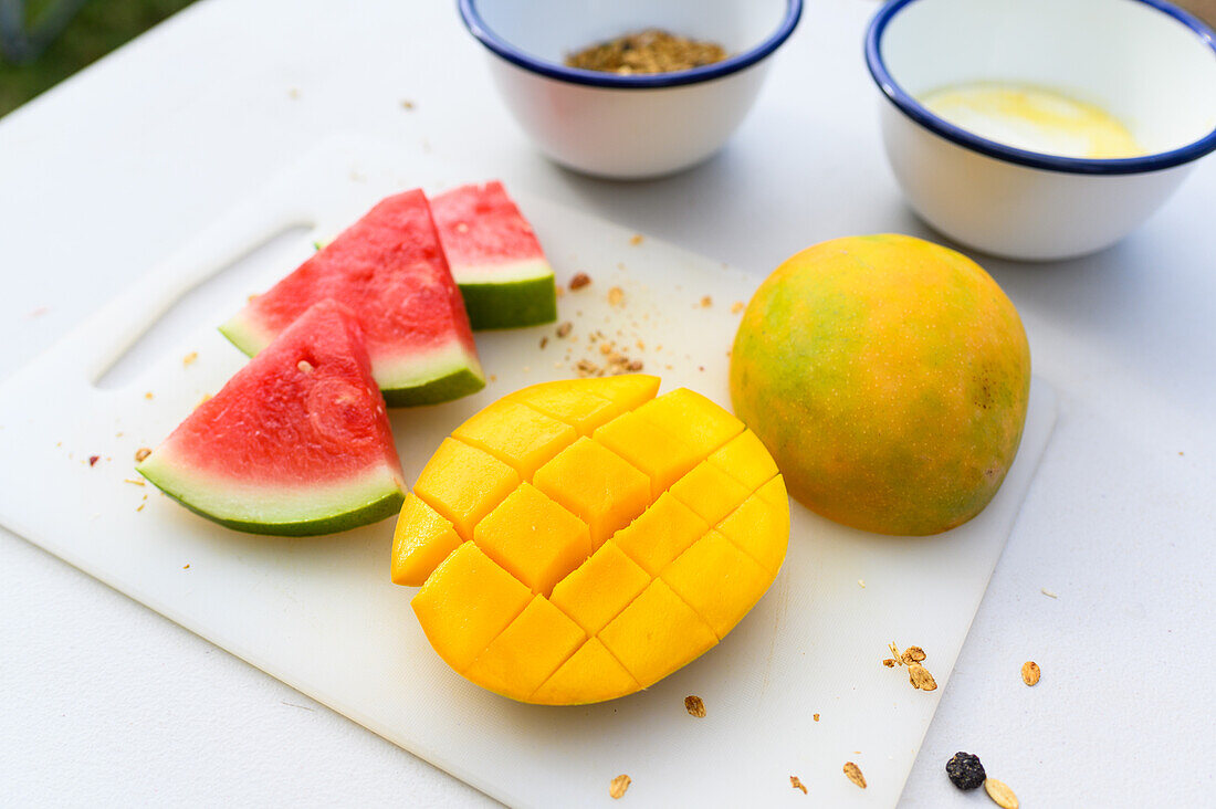 Draufsicht auf frische, in Scheiben geschnittene Wassermelone und orangefarbene, saftige, halbierte Mango, die mit einem Messer auf ein weißes Schneidebrett gelegt wird