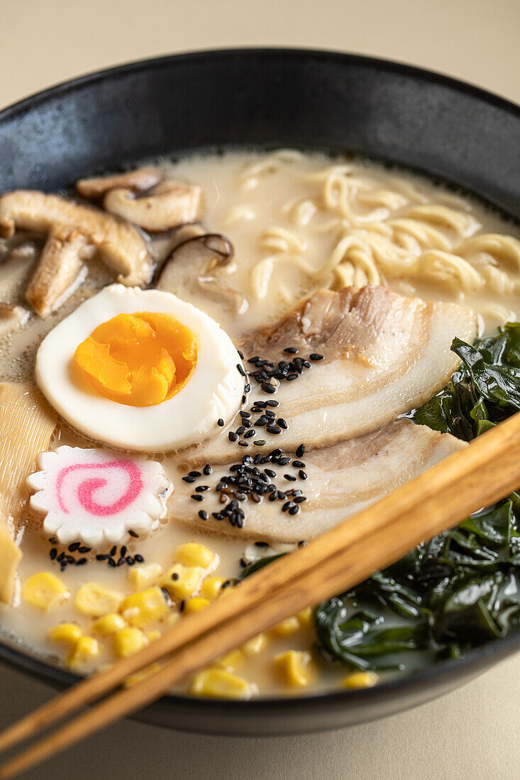 Appetitanregende japanische Ramen mit gekochtem Ei und Pilzen, serviert in einer Schüssel mit Holzstäbchen vor beigem Hintergrund