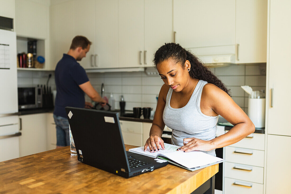 Young African American female with curly hair in home top notes in copybook while sitting at counter with laptop in kitchen with blurred man washing dishes on background